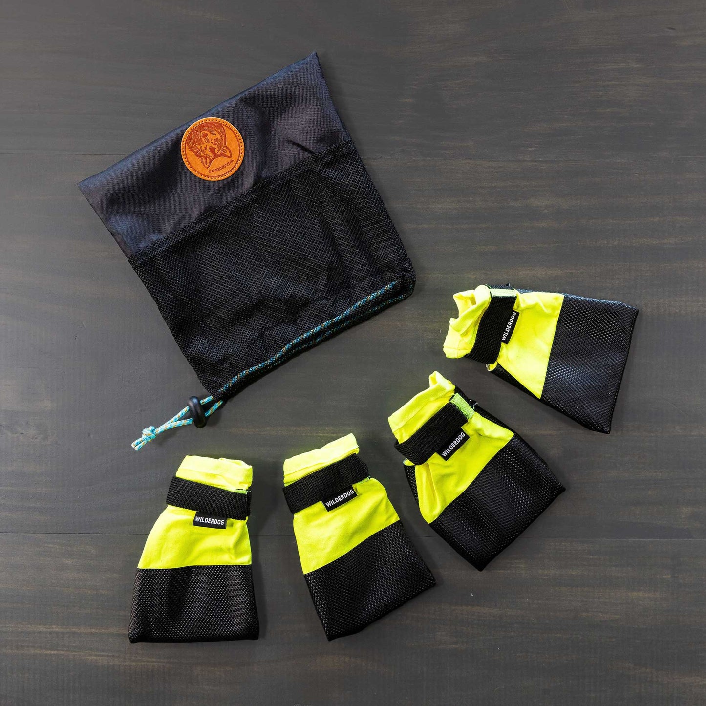 A set of Dog Booties, featuring four yellow and black booties with Velcro straps for excellent paw protection, is arranged in a semi-circle on a gray surface. Positioned above them is a black mesh storage bag with a drawstring and adorned with a brown leather patch.