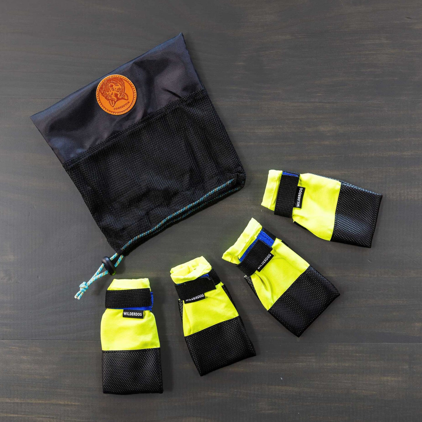 A set of four dog booties in yellow and black, featuring Velcro straps for paw protection, is placed atop a dark surface. Close by is a black mesh drawstring pouch that boasts a brown leather patch, suggesting the use of breathable fabric in its construction.