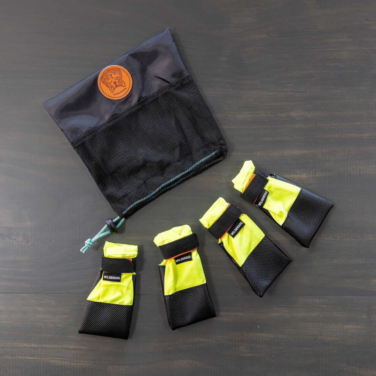 A set of Dog Booties featuring four black and yellow protective paw wear with reflective stripes, secure labeled Velcro straps, presented in a black mesh drawstring bag with a brown logo, displayed on a dark surface for sled dog racers.