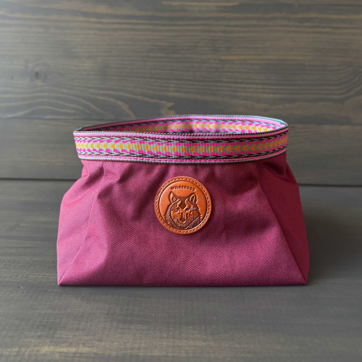 A maroon fabric bag with colorful woven trim and a round leather patch embossed with a design makes the perfect carrier for your Backpacking Bowl. Set against a wooden background, it's ideal for any adventure.