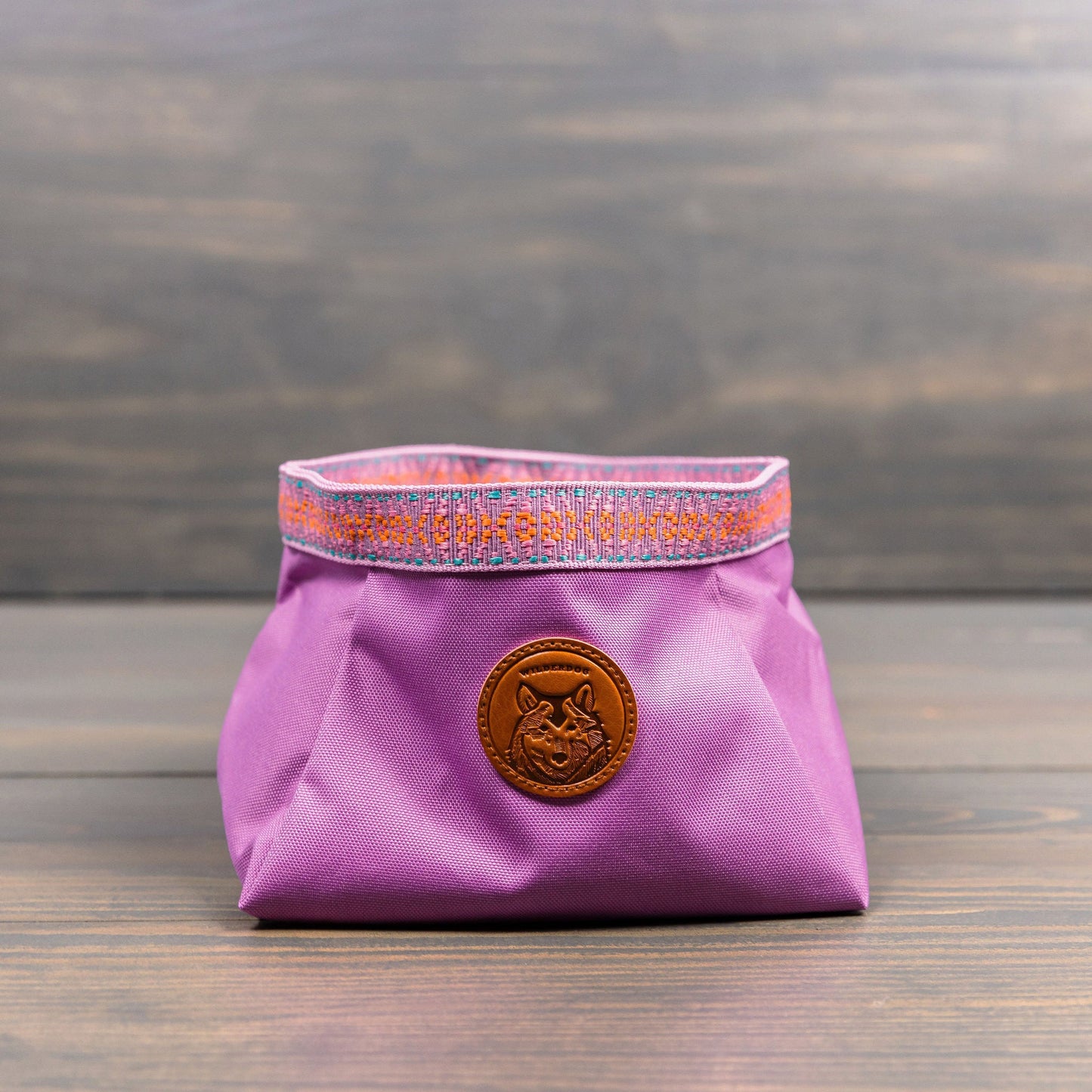 A compact purple "Backpacking Bowl" made of fabric, featuring a colorful patterned trim at the top and adorned with a distinctive round brown leather patch on the front. This water-resistant accessory is perfect for carrying essentials during outdoor adventures and is displayed on a wooden surface against a blurred wood background.