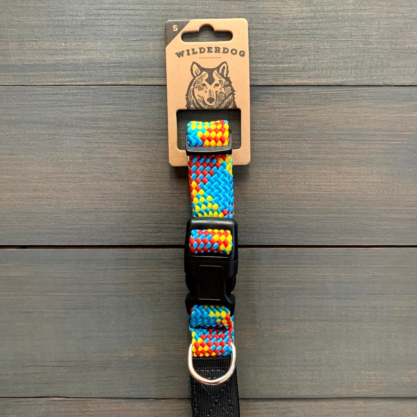 The Aprés Collar, a vibrant and durable dog collar adorned with a woven rainbow pattern, is showcased on a wooden surface. It features a black plastic buckle and a metal ring. The packaging displays the brand name "Wilderdog" along with an illustration of a dog on the label.