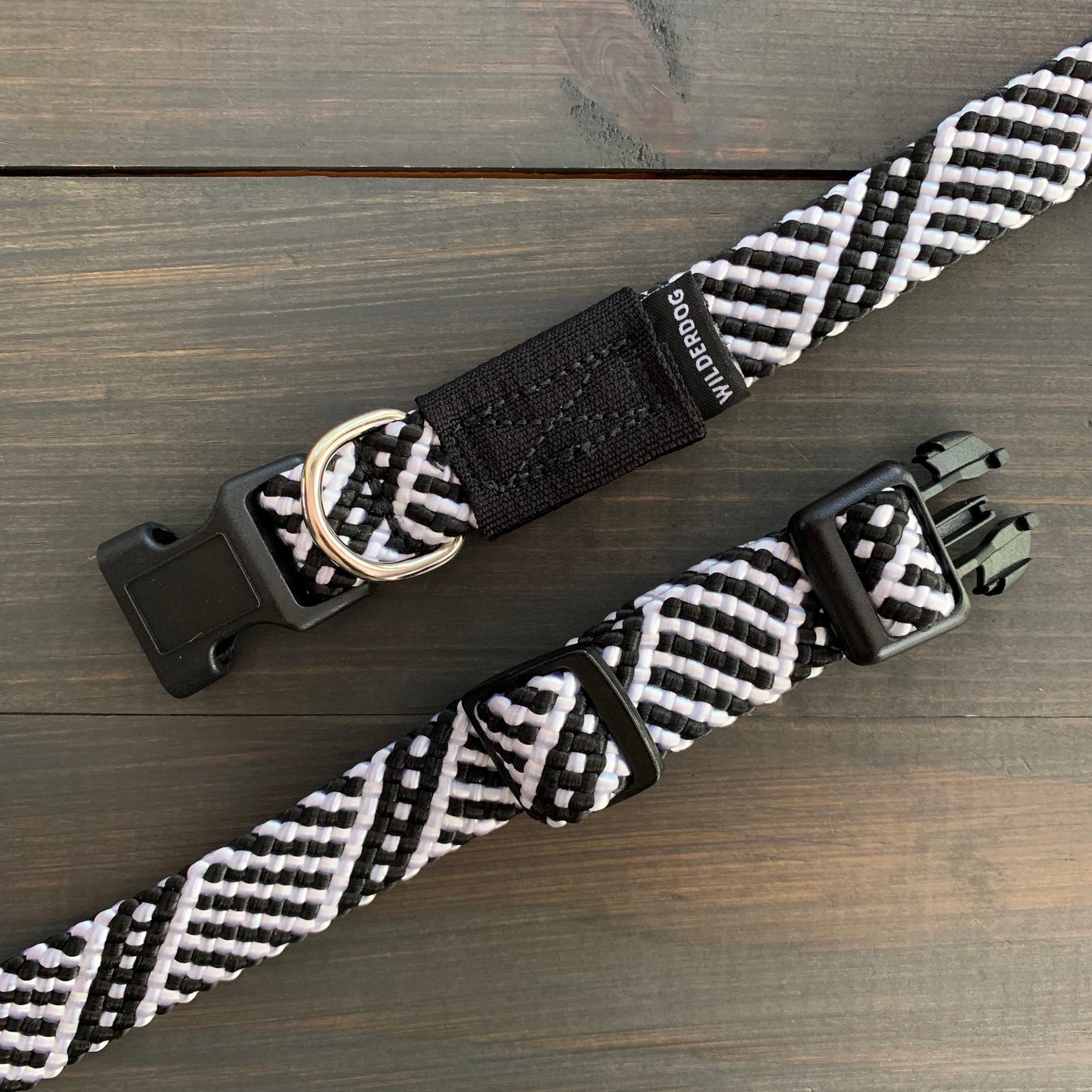 Close-up of two Black and White Collars, resembling black and white woven rock climbing rope, with plastic buckles and metal D-rings, placed on a dark wooden surface. The collars feature interlacing patterns and a small label with text partially visible.