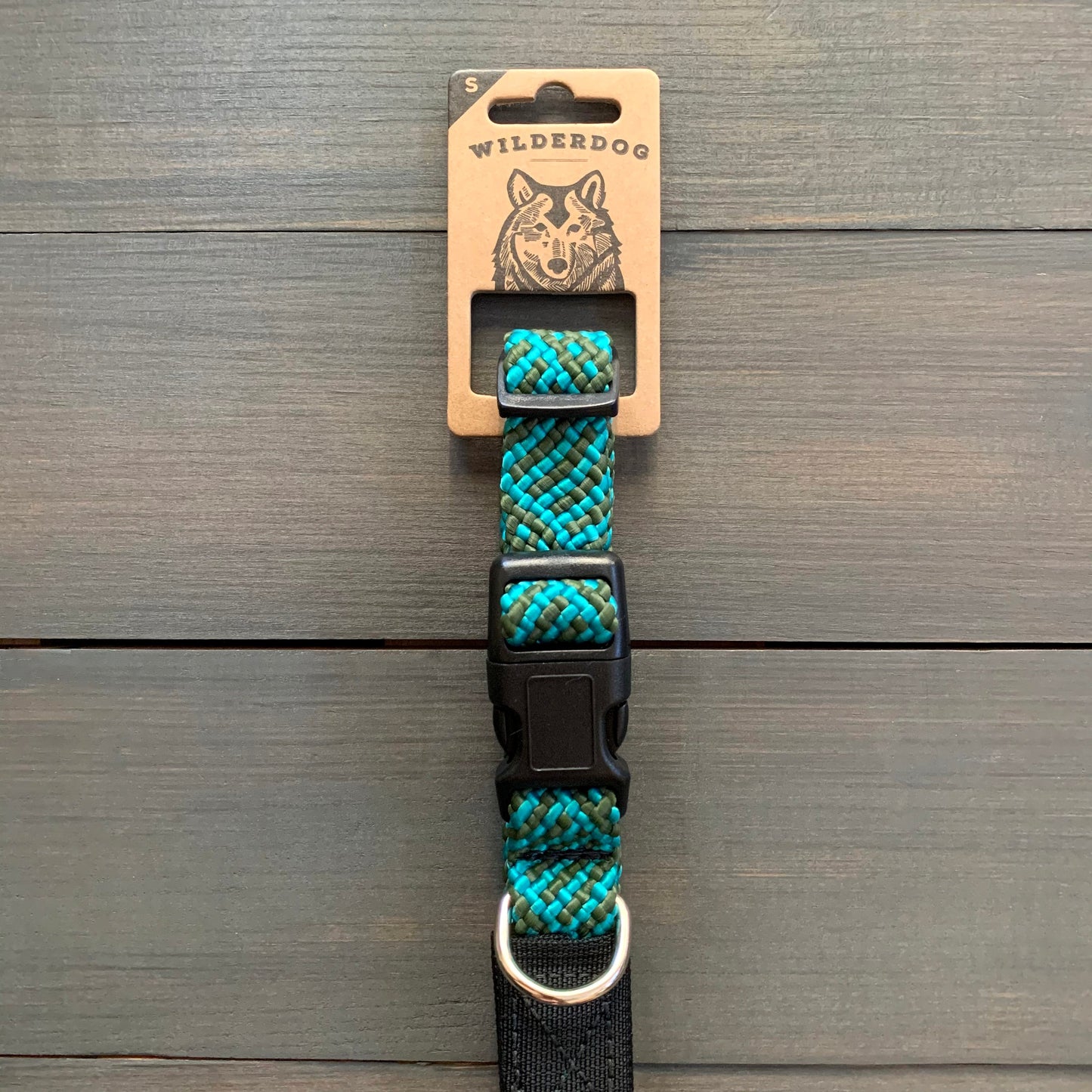 The Cascade Collar, a teal and black patterned dog collar made from durable rock climbing rope, features a black plastic buckle and metal D-ring. Displayed on a cardboard packaging label showcasing the "Wilderdog" logo, it proudly highlights its lifetime warranty. The collar is set against a wooden background.