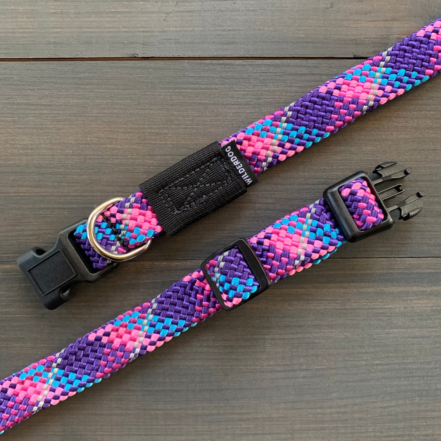The Razzleberry Reflective Collar, known for its durability and vibrant pattern of purple, blue, pink, and white, is displayed. It features a black plastic buckle, a size-adjustment slider, and a metal D-ring for attaching a leash or tags. The collar is shown laid flat on a wooden surface and comes with a lifetime warranty.