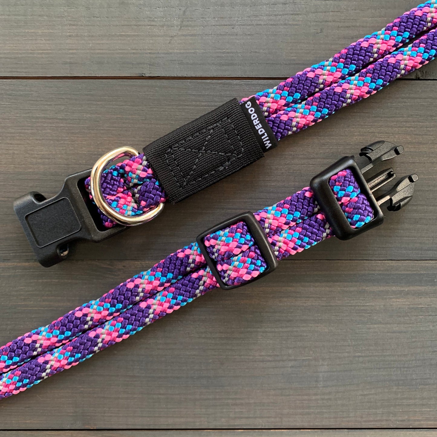 The Razzleberry Reflective Collar is a durable dog collar with a vibrant, multicolored design featuring shades of purple, pink, and blue. It includes a black plastic buckle, a metal D-ring for attaching a leash, and an adjustable strap. Covered by a lifetime warranty, the collar is laid out on a dark wooden surface.