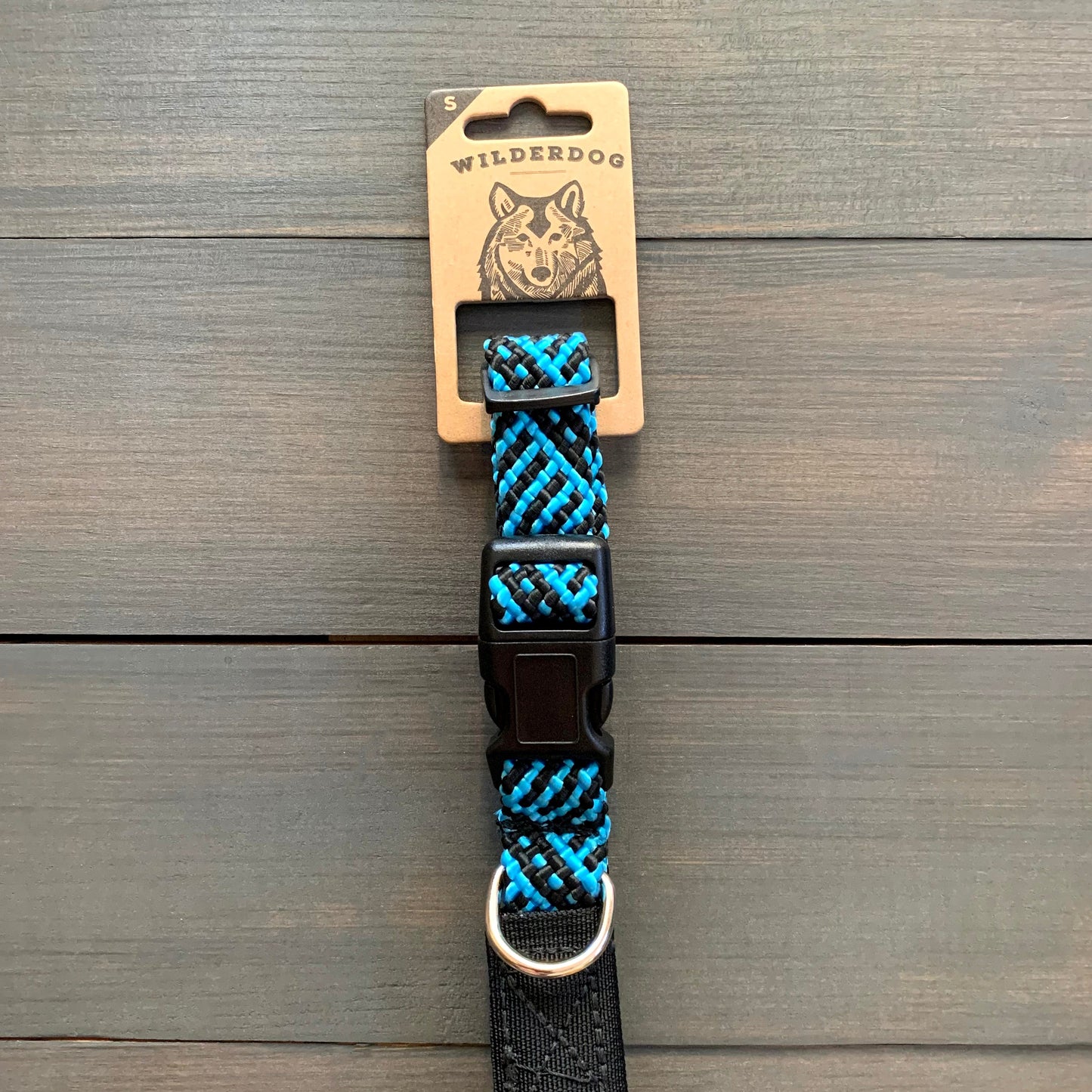 A Teton Collar by Wilderdog, in blue and black woven hues, crafted with the durability of rock climbing rope, displayed on a wooden surface. The packaging features a dog illustration. The collar includes a black buckle and a metal D-ring for attaching a leash or tag.