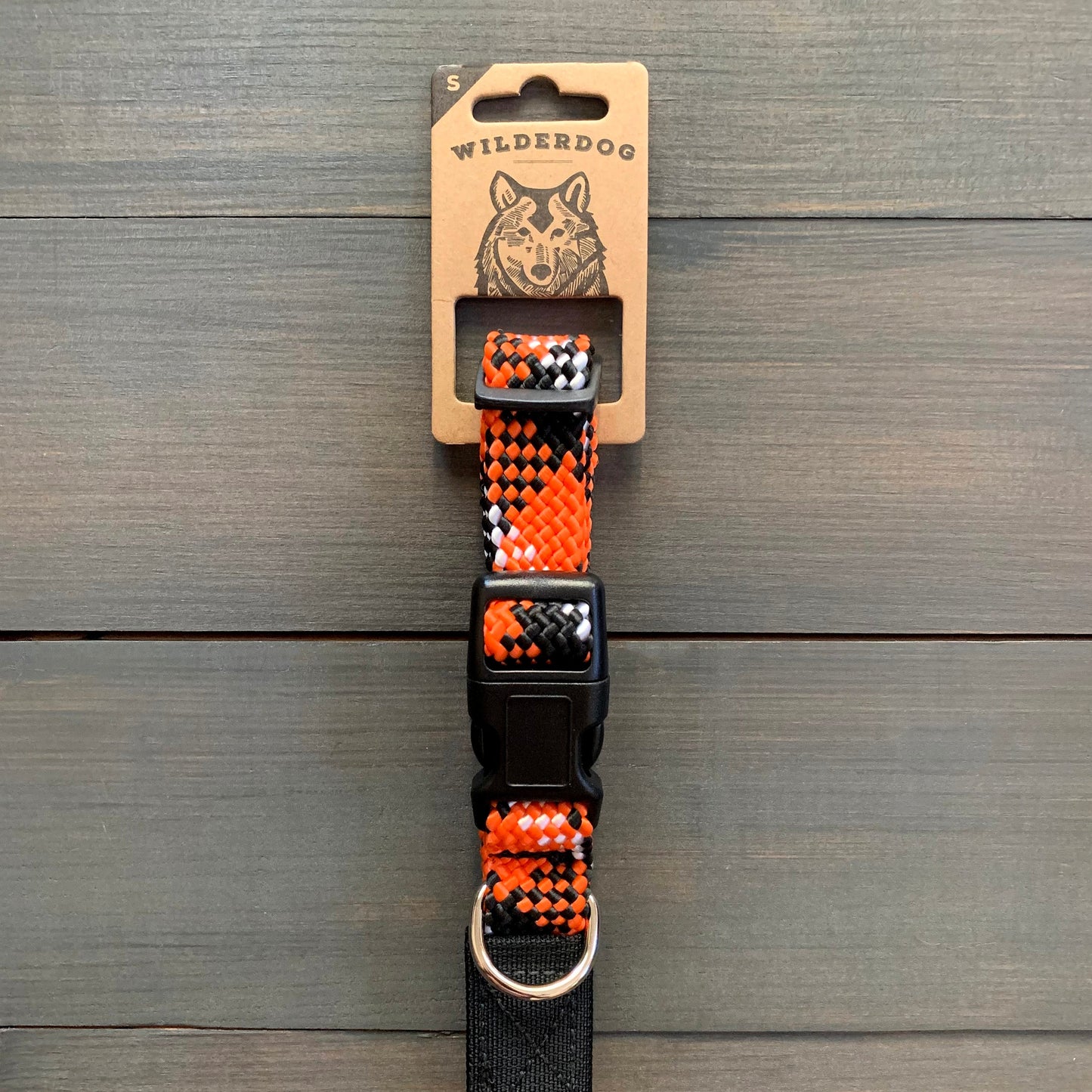The Wildcat Collar, a vibrant and heavy-duty dog collar featuring an orange, black, and white woven pattern, is displayed against a wooden background. The collar is packaged on a cardboard tag labeled "Wilderdog," showcasing an illustration of a dog's face. A black plastic buckle and metal ring are also visible.