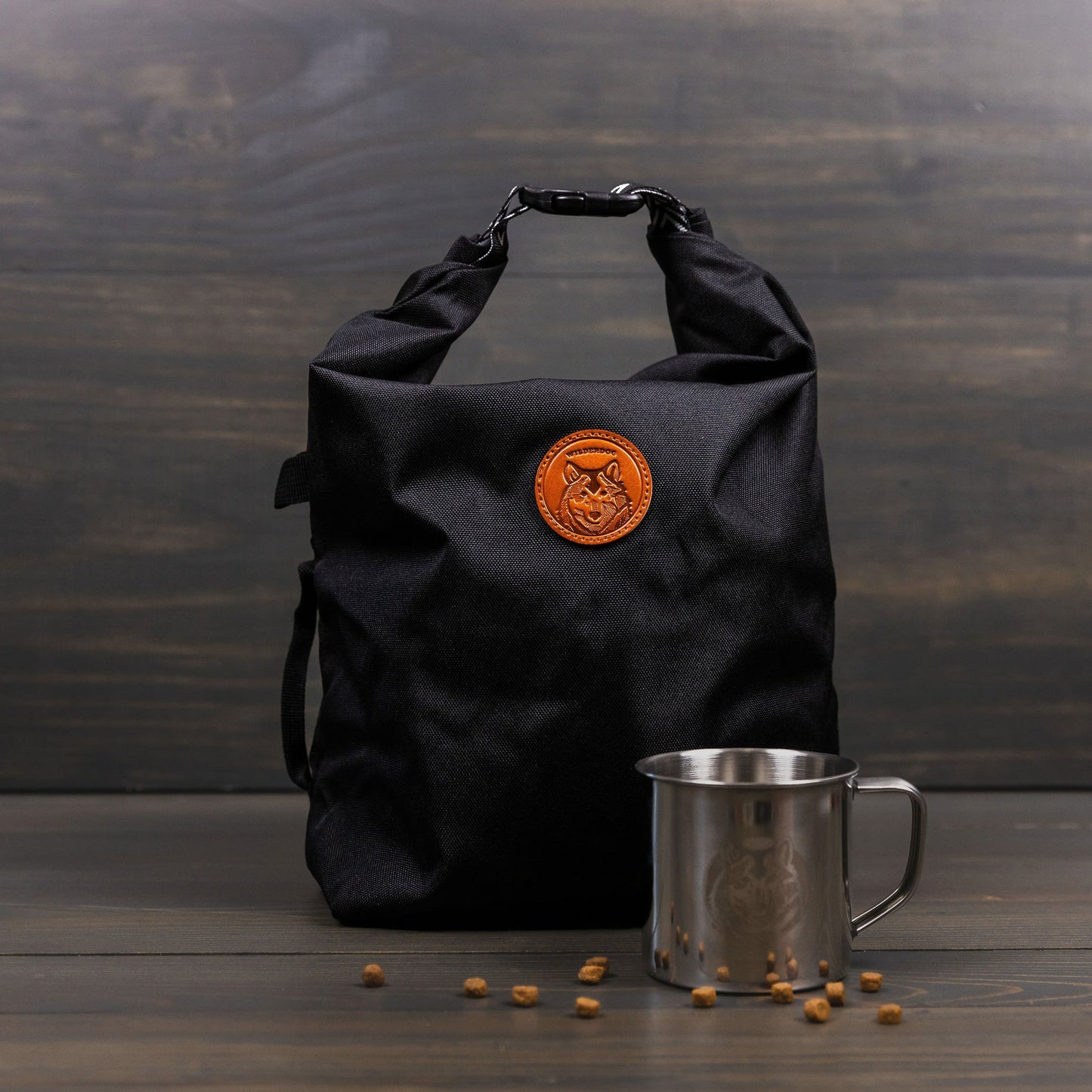 A black roll-top Doggie Bag, featuring a brown leather patch logo, is elegantly positioned on a wooden surface, combining sophistication with practicality. Adjacent to it lies a stainless steel cup surrounded by scattered coffee beans, all set against the rich background of a dark wooden wall.