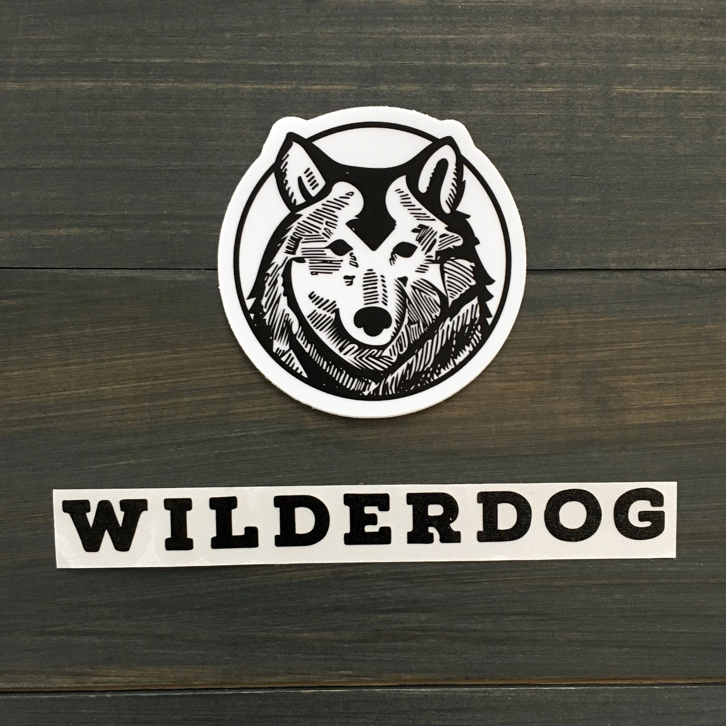 A black and white waterproof vinyl decal featuring a detailed illustration of a wolf's head inside a circle is placed on a dark wooden surface. Below the Wilderdog Sticker Decals, the word "WILDERDOG" is written in bold, black, uppercase letters.