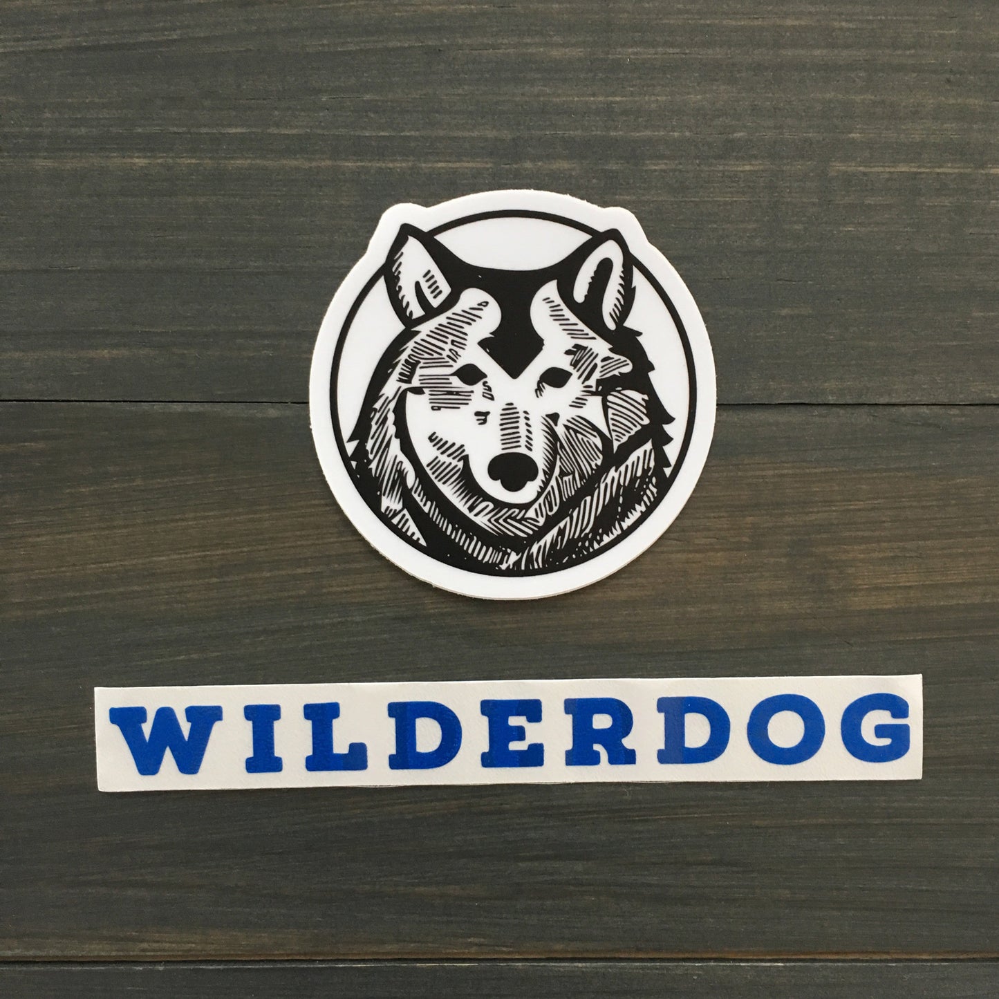 A circular logo showcases a black and white graphic illustration of a wolf's head. Below the logo, "WILDERDOG" is prominently displayed in bold blue capital letters. The background is a dark wooden surface, ideal for highlighting the durability of these outdoor-rated Wilderdog Sticker Decals.