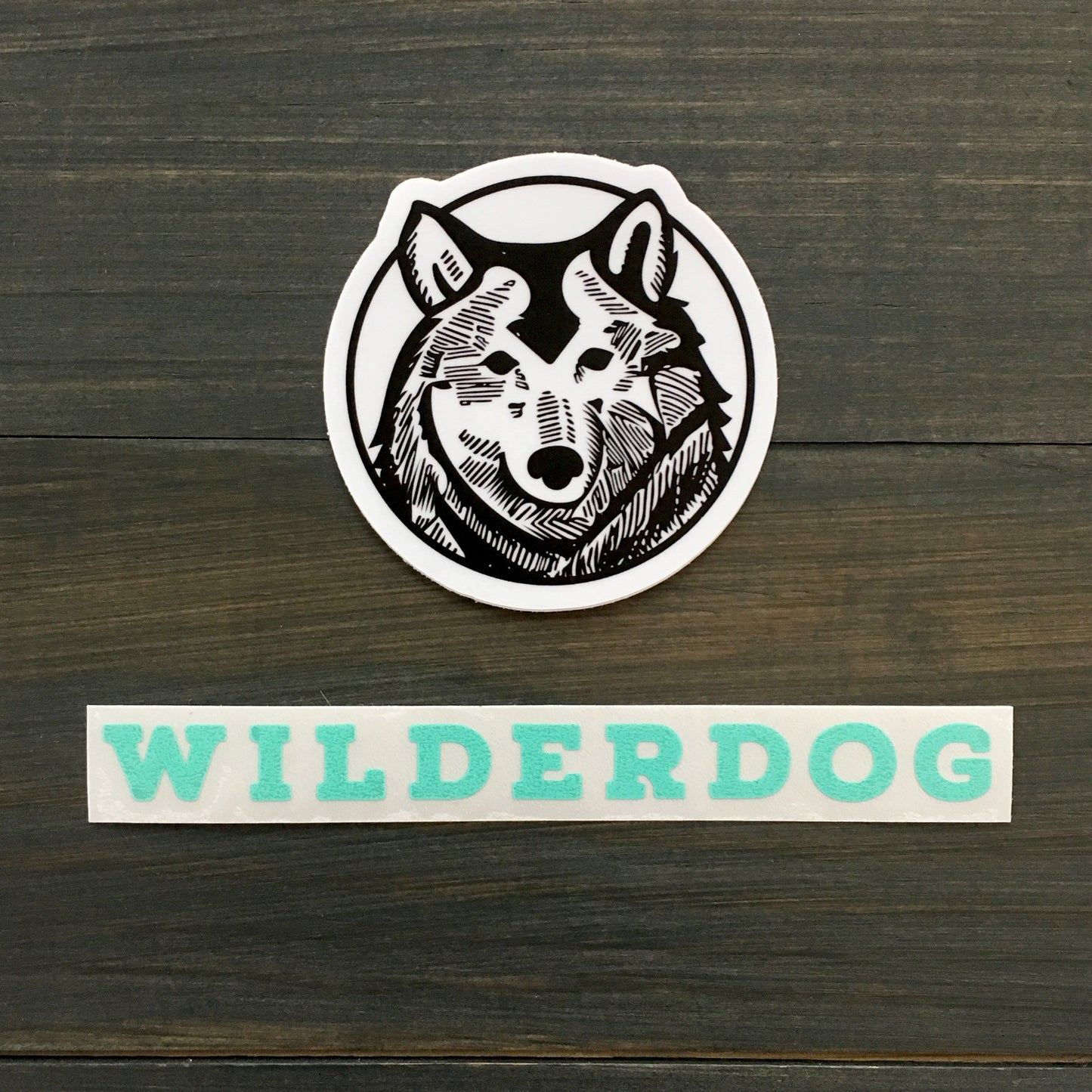 A black and white Wilderdog Sticker Decal showcases a stylized wolf's head inside a circle. Below the decal, the word "WILDERDOG" appears in bold turquoise uppercase letters. These outdoor-rated decals are set against a background of dark, wooden planks.