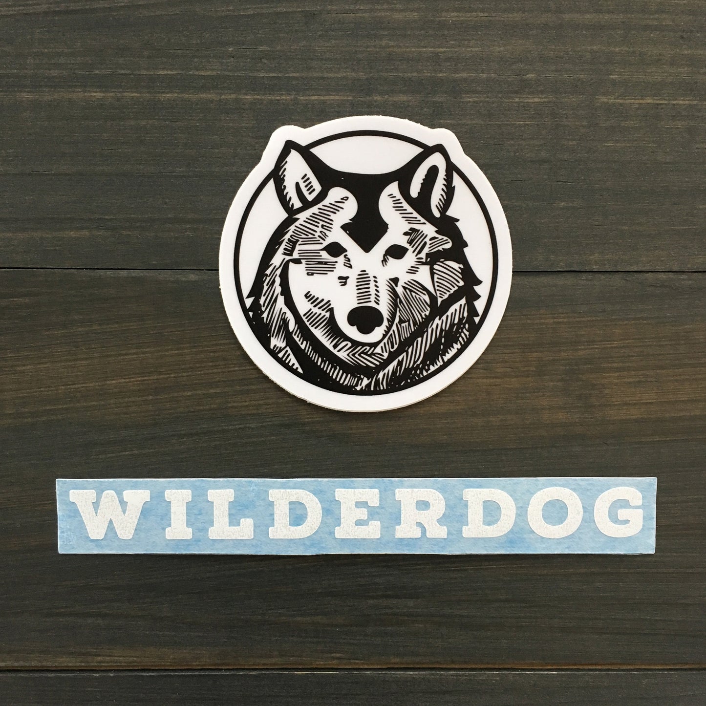 A circular Wilderdog Sticker Decal features an intricate black and white drawing of a wolf's head against a white background. Below the decal, "WILDERDOG" is boldly displayed in white letters on a light blue background. These outdoor-rated decals are placed on a dark wooden surface, making them perfect for any adventure.