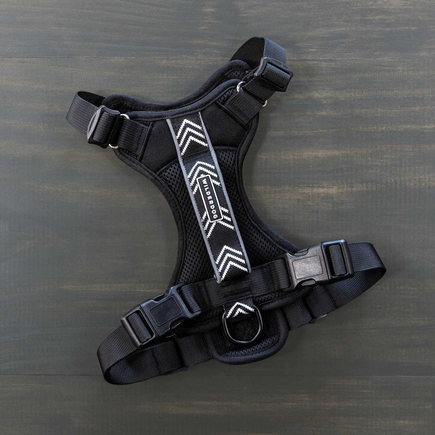 A Dog Harness in black, featuring adjustable straps and buckle closures, is laid flat on a dark wooden surface. Perfect for outdoor adventures, it boasts a pattern of white chevrons down the center, made with reflective fabric for enhanced visibility, and includes a metal ring for leash attachment.