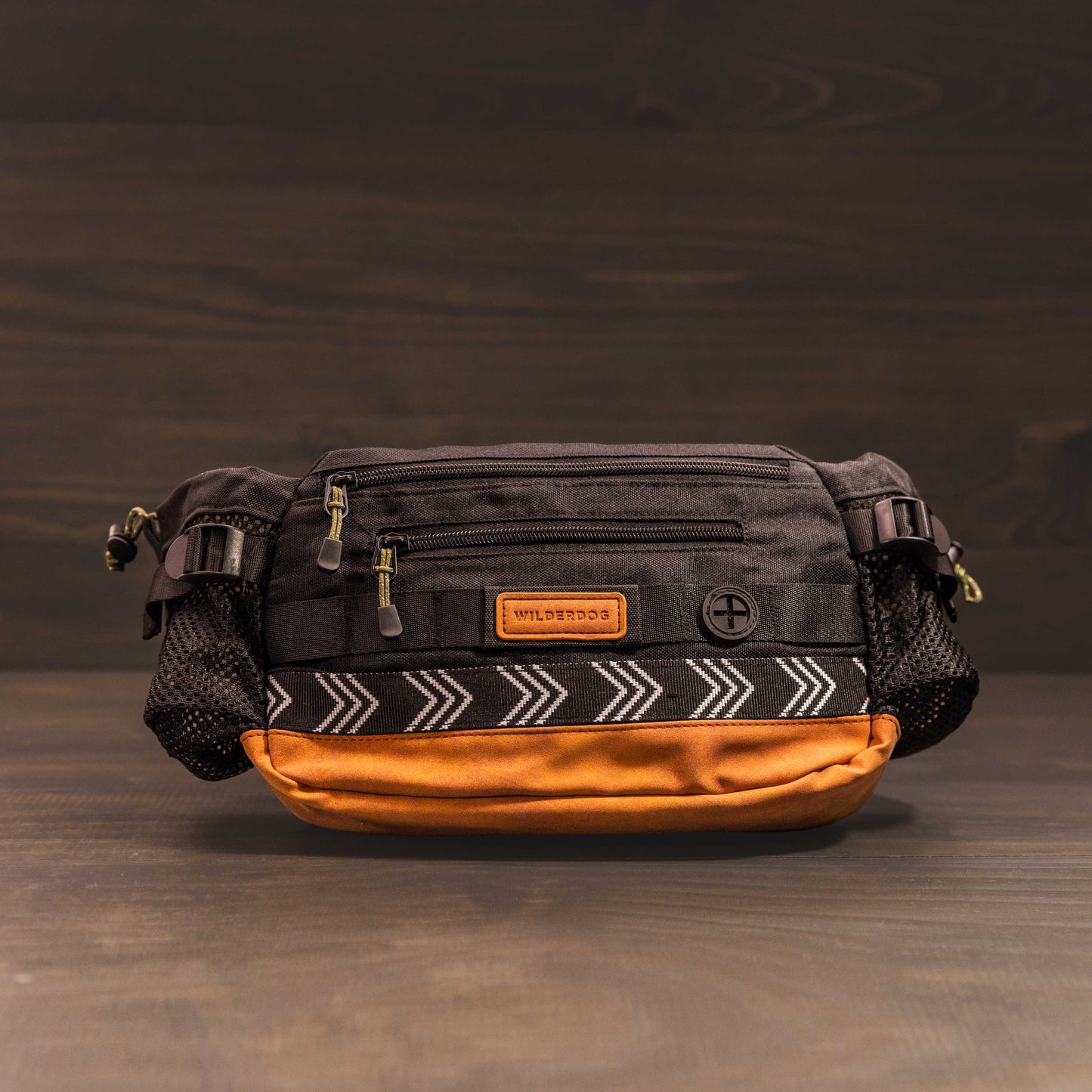 The Mega Hip Pack, available in a stylish black and brown color with patterned accents, offers a 3-liter capacity. It features multiple zippered compartments, an adjustable strap, and a convenient mesh side pocket. The pack is beautifully displayed against a dark wooden background.