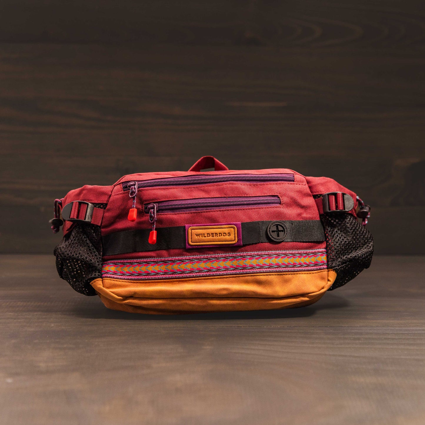 A vibrant Mega Hip Pack is showcased against a dark backdrop, displaying a mix of red, orange, and purple hues. It includes zippered compartments and mesh pockets on its sides for convenience. The adjustable strap offers a customizable fit, and the front proudly bears a "WILDBRDS" patch.