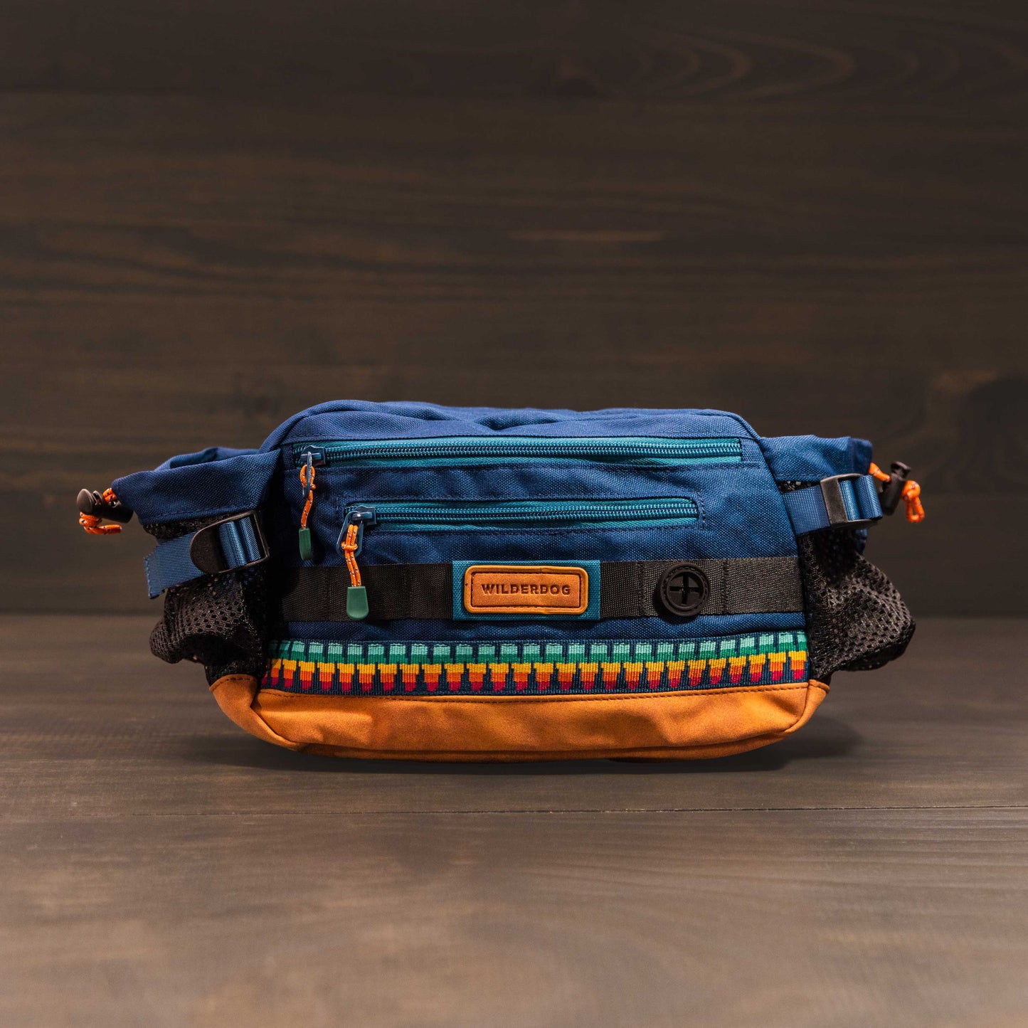Introducing the Mega Hip Pack, featuring a vibrant blue top and an eye-catching orange base. It includes black side mesh pockets for added utility, a front zip pocket, and stylish patterned trim. The pack is complemented by an adjustable strap and showcased against a wooden background.