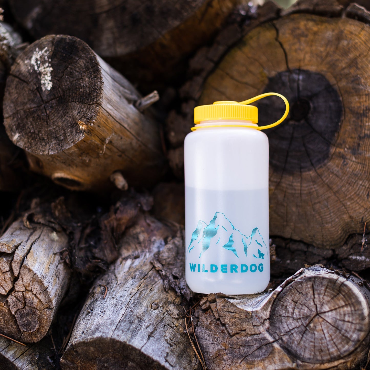 A 32oz Wilderdog Nalgene water bottle, equipped with a yellow cap and adorned with a blue "Wilderdog" logo featuring mountains, is placed against a stack of cut logs. The background showcases rough, textured wood in various shades of brown. This BPA/BPS-free bottle also includes a convenient carrying loop attached to the cap.