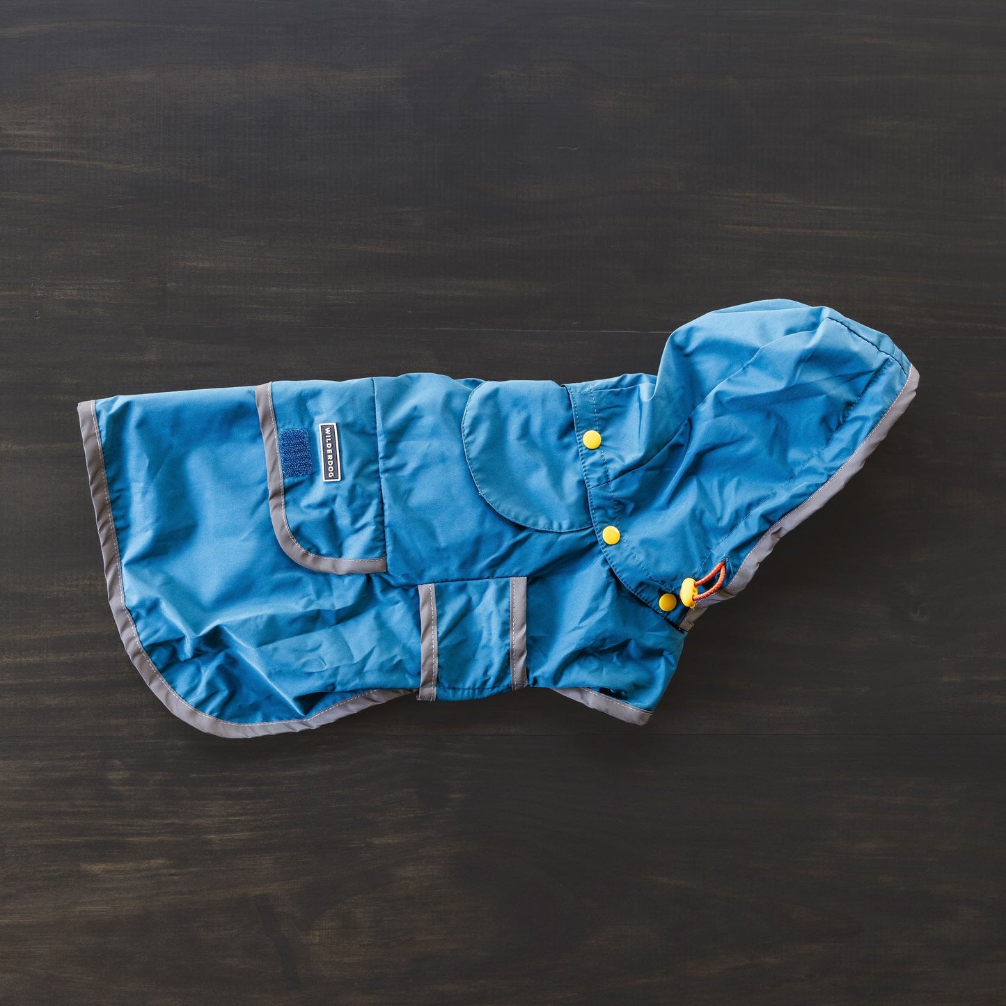 A Dog Rain Jacket in blue, designed to be waterproof and featuring a hood with reflective trim, is spread out on a dark wooden surface. The jacket includes pockets, a breathable mesh lining, and yellow snap buttons for closure.
