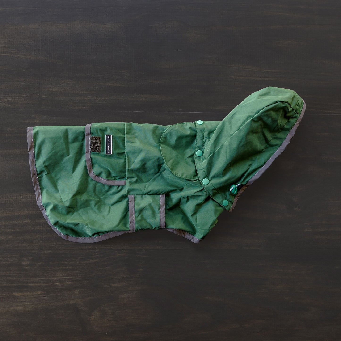 Reflective trim accents the green Dog Rain Jacket, which features a hood and pockets, and is laid flat on a dark wooden surface. This waterproof jacket also includes a breathable mesh interior, ensuring comfort and safety for your furry friend in all weather conditions.