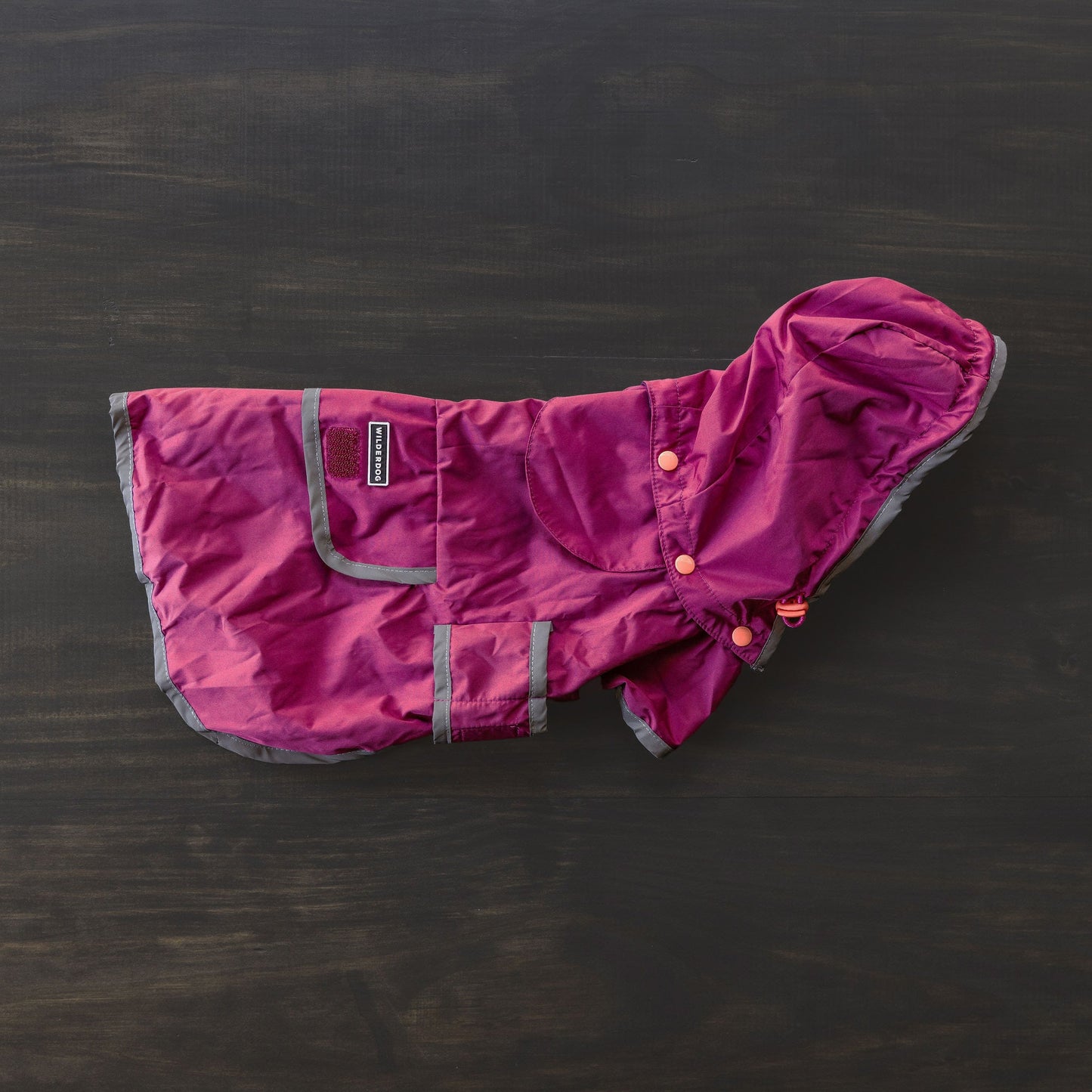 The Dog Rain Jacket, featuring a maroon and gray waterproof design with a hood and snap buttons, is laid flat on a dark surface. It includes a breathable mesh interior, reflective trim, a small brand label on the pocket, and velcro straps for securing.
