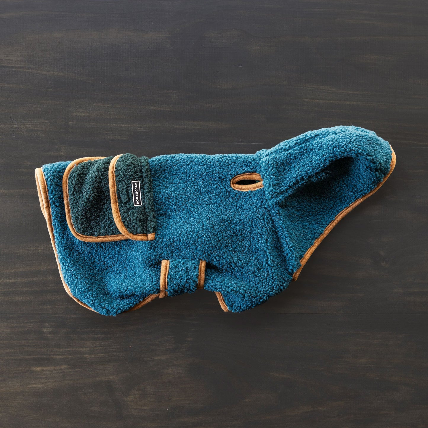 A blue Sherpa Jacket, tailored as a snug dog coat, is spread out on a dark wooden surface. It includes a hood, tan trim, adjustable side straps for a perfect fit, and a convenient leash hole for effortless outdoor adventures.