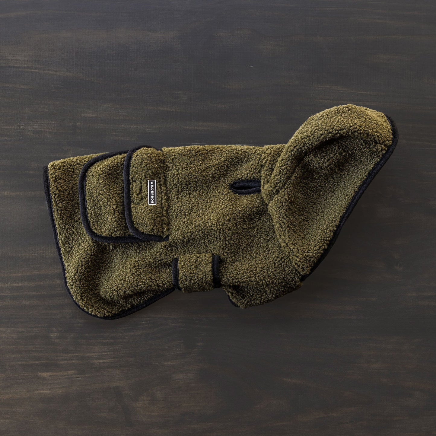 The Sherpa Jacket, a green fleece dog coat with black trim and adjustable straps, is displayed on a dark wooden surface. This cozy jacket includes a small leash hole for added convenience.