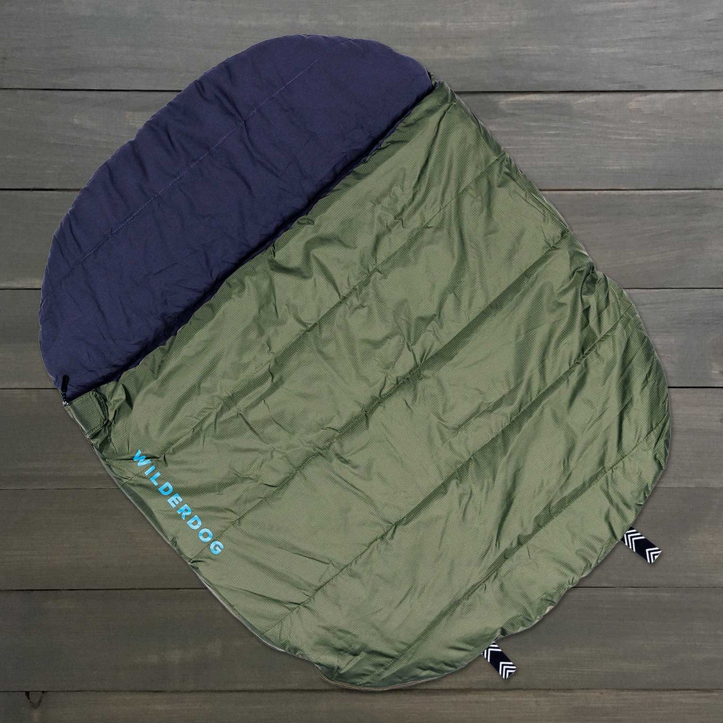 A green and navy "Wilderdog" sleeping bag in a rectangular quilted design rests on a wooden surface. Made from durable ripstop polyester, it's ideal for indoor or outdoor adventures with your pet.