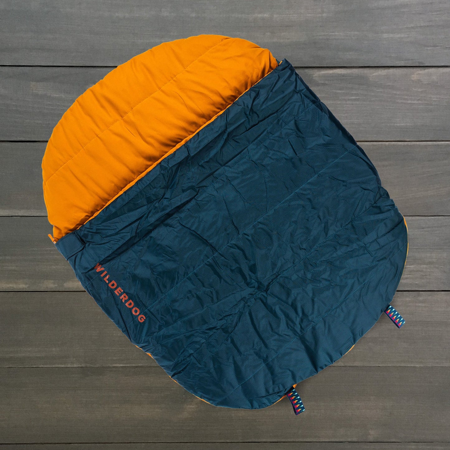 A two-toned WILDERDOG sleeping bag, made of durable polyester with an orange top and dark blue bottom, rests on a wooden floor. It has colorful cords at the bottom corners, perfect for outdoor adventures.