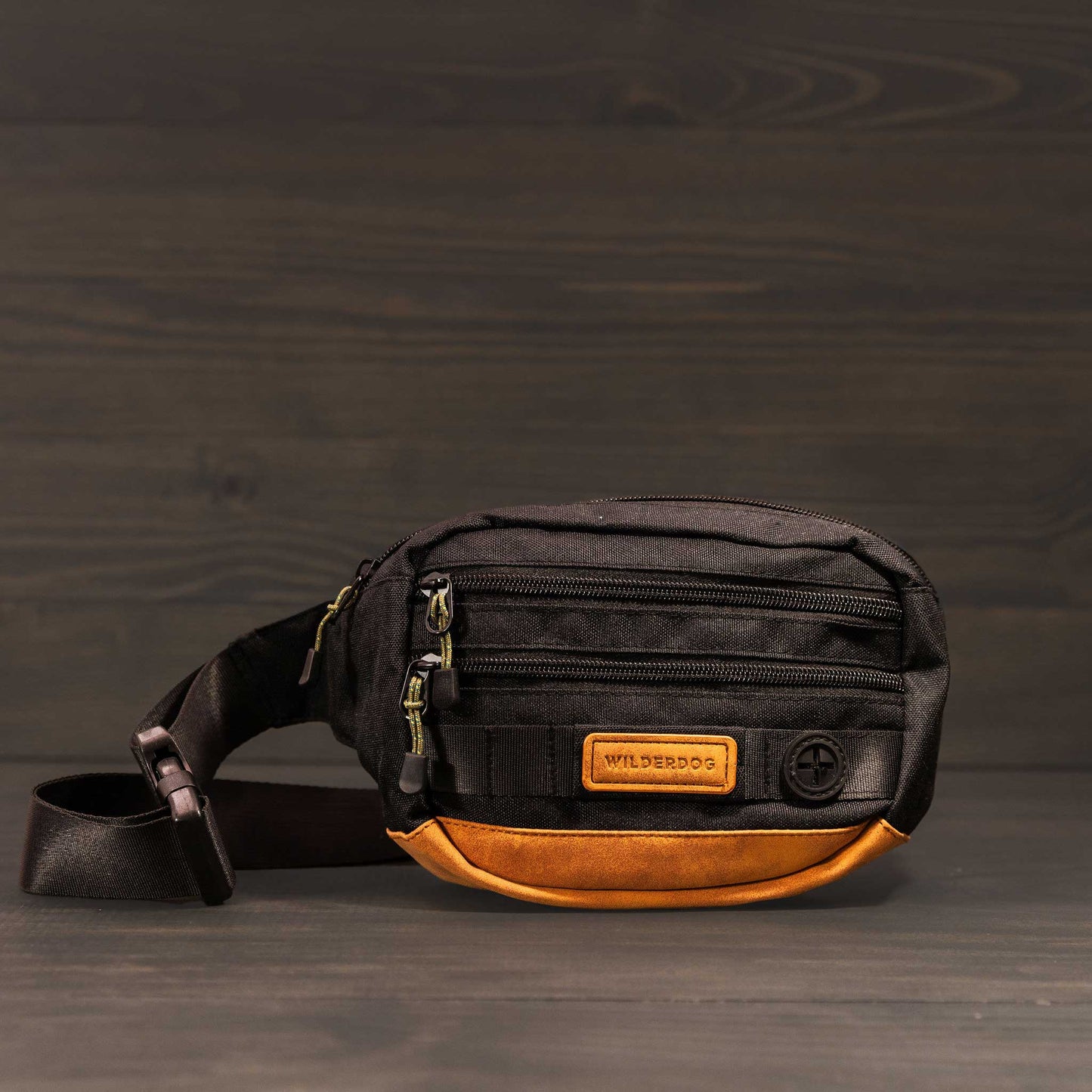 A black and brown Utility Pack by Wilderdog, featuring multiple zippered compartments and crafted from vegan material with a black adjustable strap, displayed against a dark wooden background.