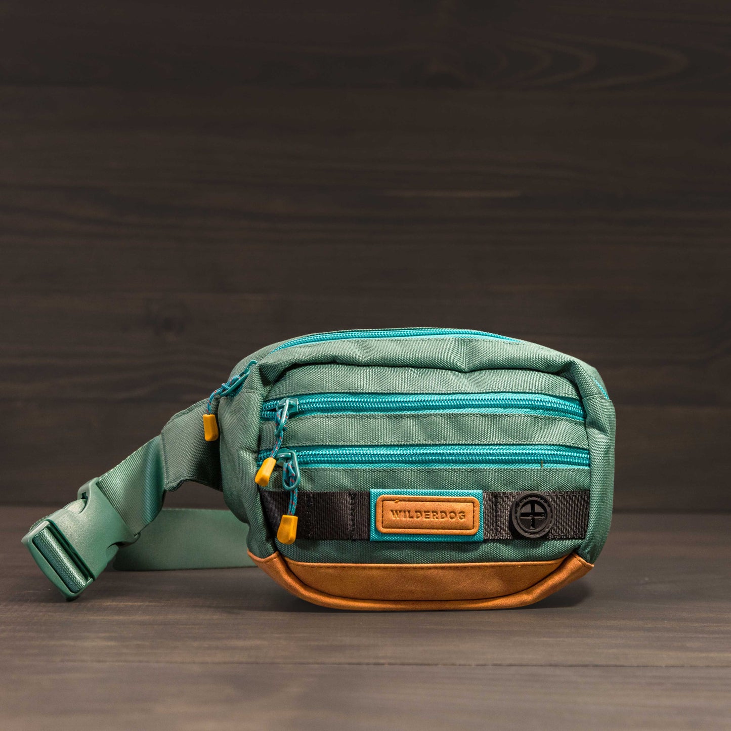 The Utility Pack, crafted from vegan material in green and brown, showcases two blue-zippered compartments and a beige "Wilderdog" label set against a dark wooden background. This treat bag is designed with a side strap and small yellow zipper pulls.