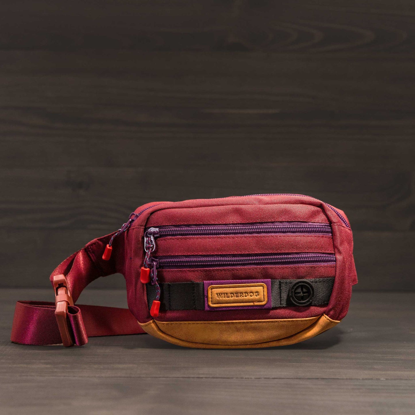 Introducing the Utility Pack: a compact and chic pouch in maroon and orange fabric, made from vegan materials. It features multiple zippered compartments and an adjustable strap, showcased against a dark, textured surface.