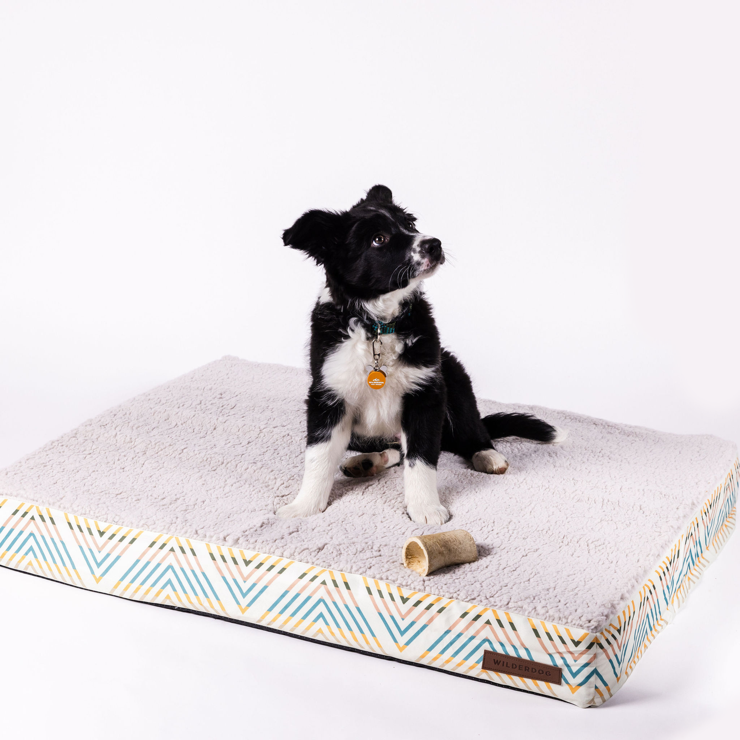 A small black and white dog sits on a large Memory Foam Dog Bed with a soft, textured surface. The washable cover boasts a blue and white chevron patterned border with yellow accents. Two small dog toys lie next to the dog on the non-skid bottom of the bed. The background is white.