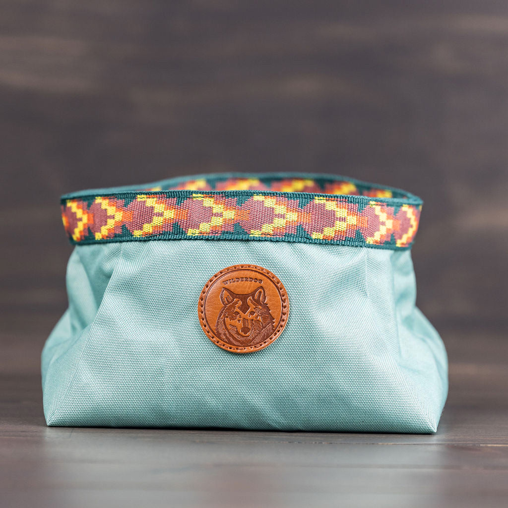 A light blue fabric bag featuring a colorful, patterned strap at the top and a front round leather patch embossed with an animal design, possibly a wolf. Ideal for outdoor adventures, it pairs perfectly with the Backpacking Bowl to cater to your furry companion's needs.