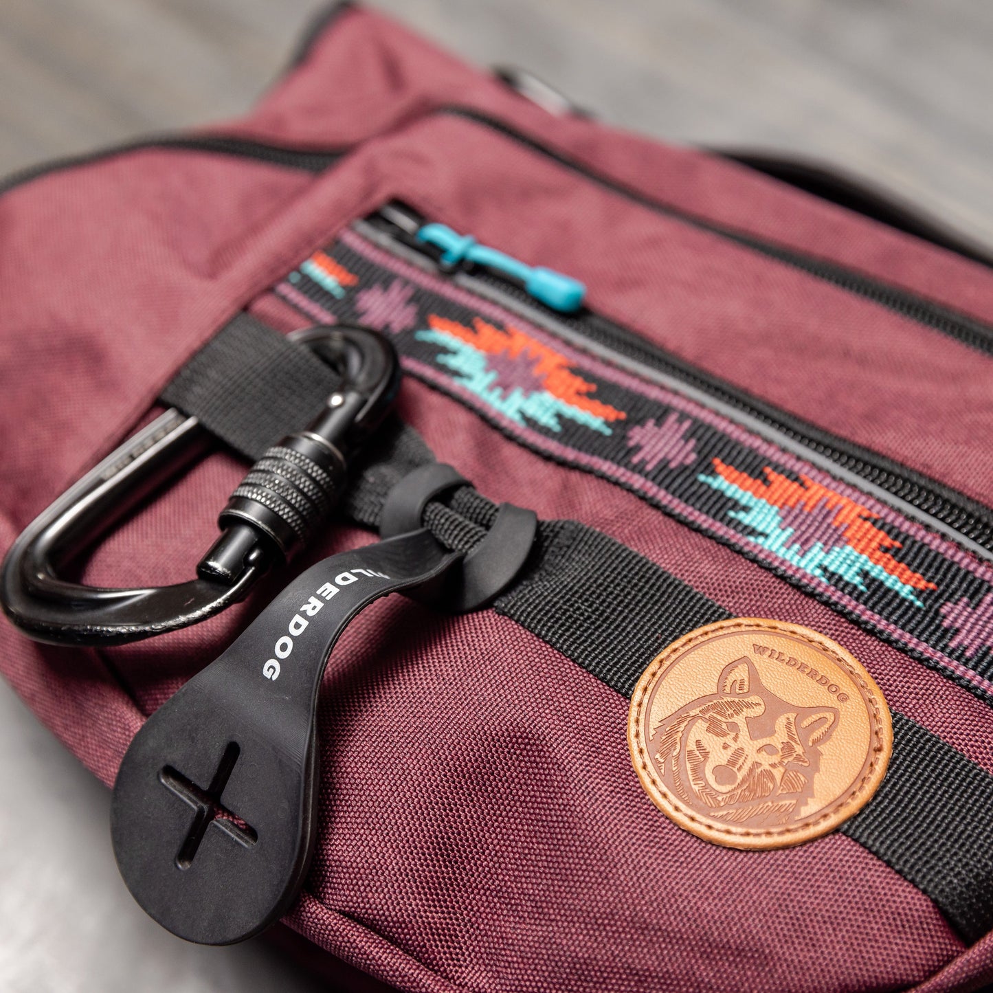 A close-up photo of the Dog Backpack in burgundy, featuring black zippers and a front pocket adorned with a colorful Native American-inspired pattern. Attached to the pocket is a black carabiner clip on a black plastic tag, complemented by a brown leather patch showcasing a dog logo.