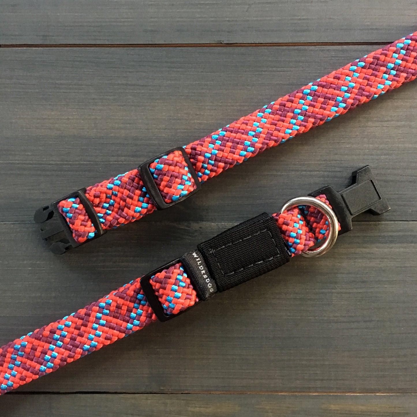 A close-up of the Maple Collar, showcasing its vibrant red, pink, blue, and white pattern on a woven fabric, rests on a wooden surface. This durable dog collar features a plastic snap buckle, an adjustable strap, and a metal D-ring for leash attachment.