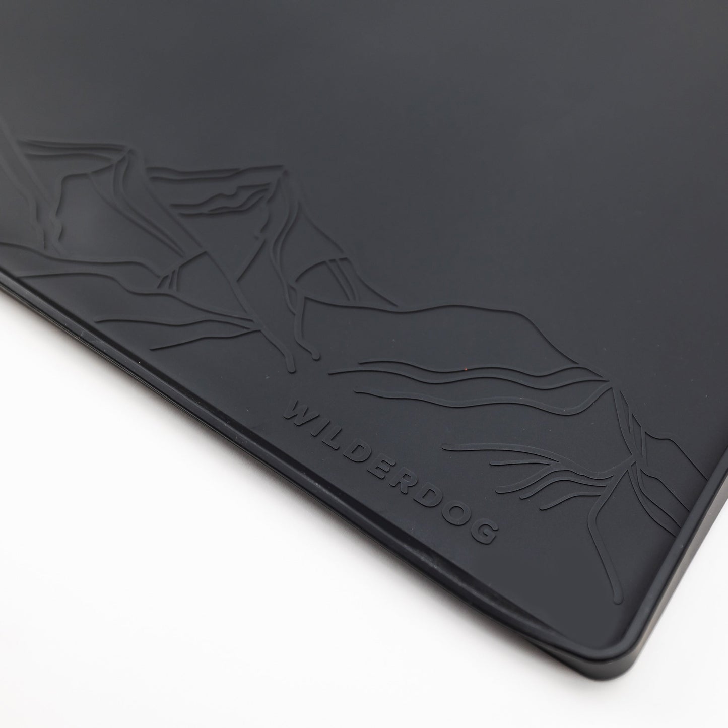 A detailed close-up of the Silicone Placemat reveals its black, rectangular form with a mountain design embossed in the top left corner. The word "WILDERDOG" is prominently embossed at the bottom center of this food-grade silicone mat. The background is white.
