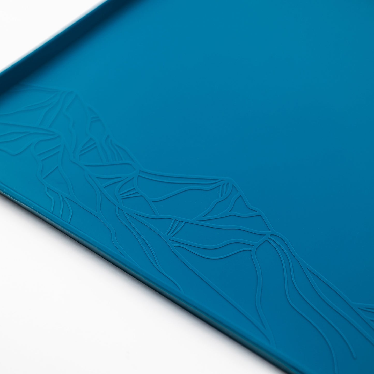 Close-up of the Silicone Placemat, featuring a rectangular, blue design made from food-grade silicone. The mat showcases an embossed pattern depicting a stylized mountain range. Set against a white background, the intricate design is highlighted in one corner of the non-slip placemat.