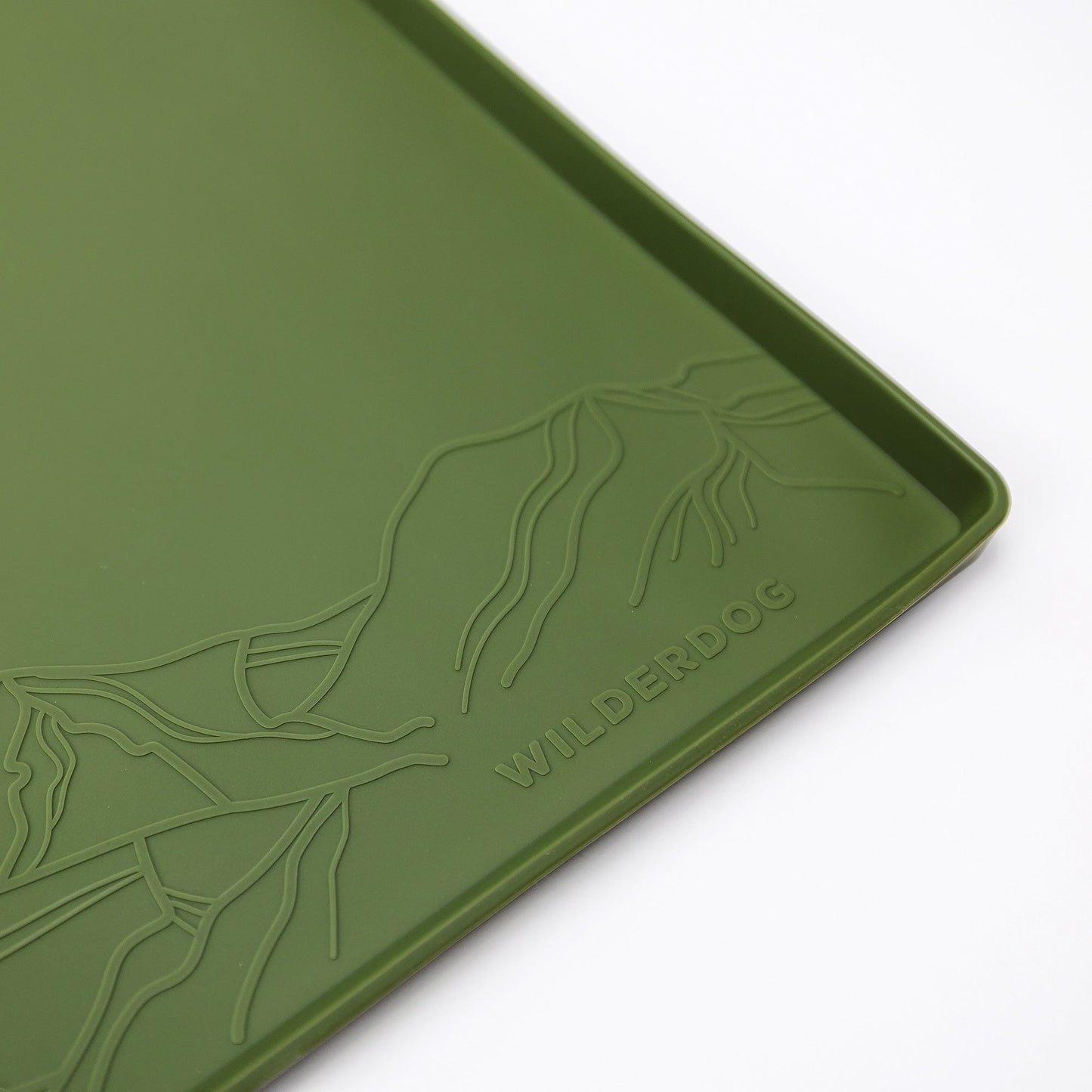 A close-up of a green Silicone Placemat by Wilderdog. Made from food-grade silicone, the mat features an embossed design of mountain outlines in one corner above the brand name "WILDERDOG" in uppercase letters. The material appears smooth with a slight texture and is non-slip.