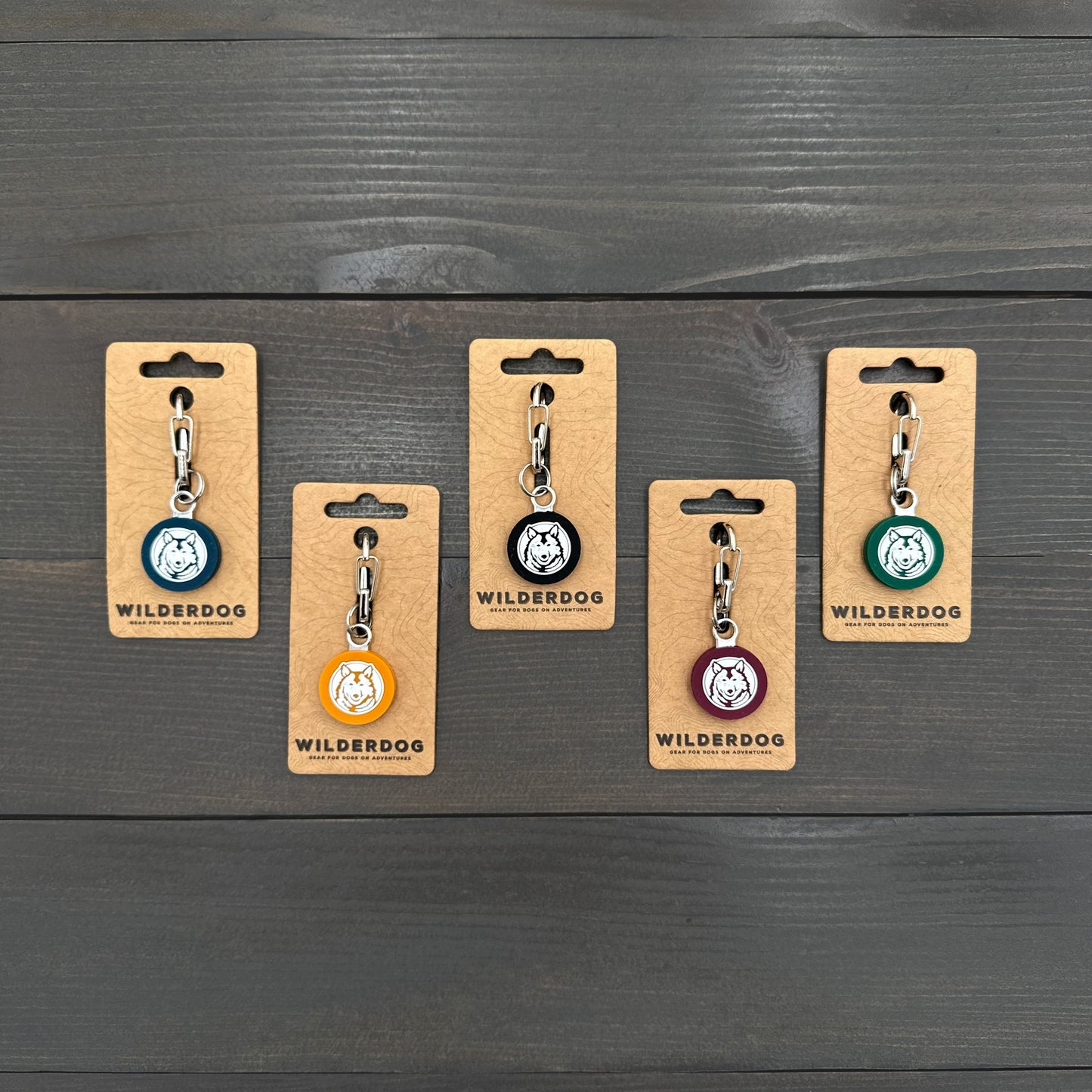 Five Wilderdog Silicone Tags in various colors with mini carabiners on a dark wooden surface.