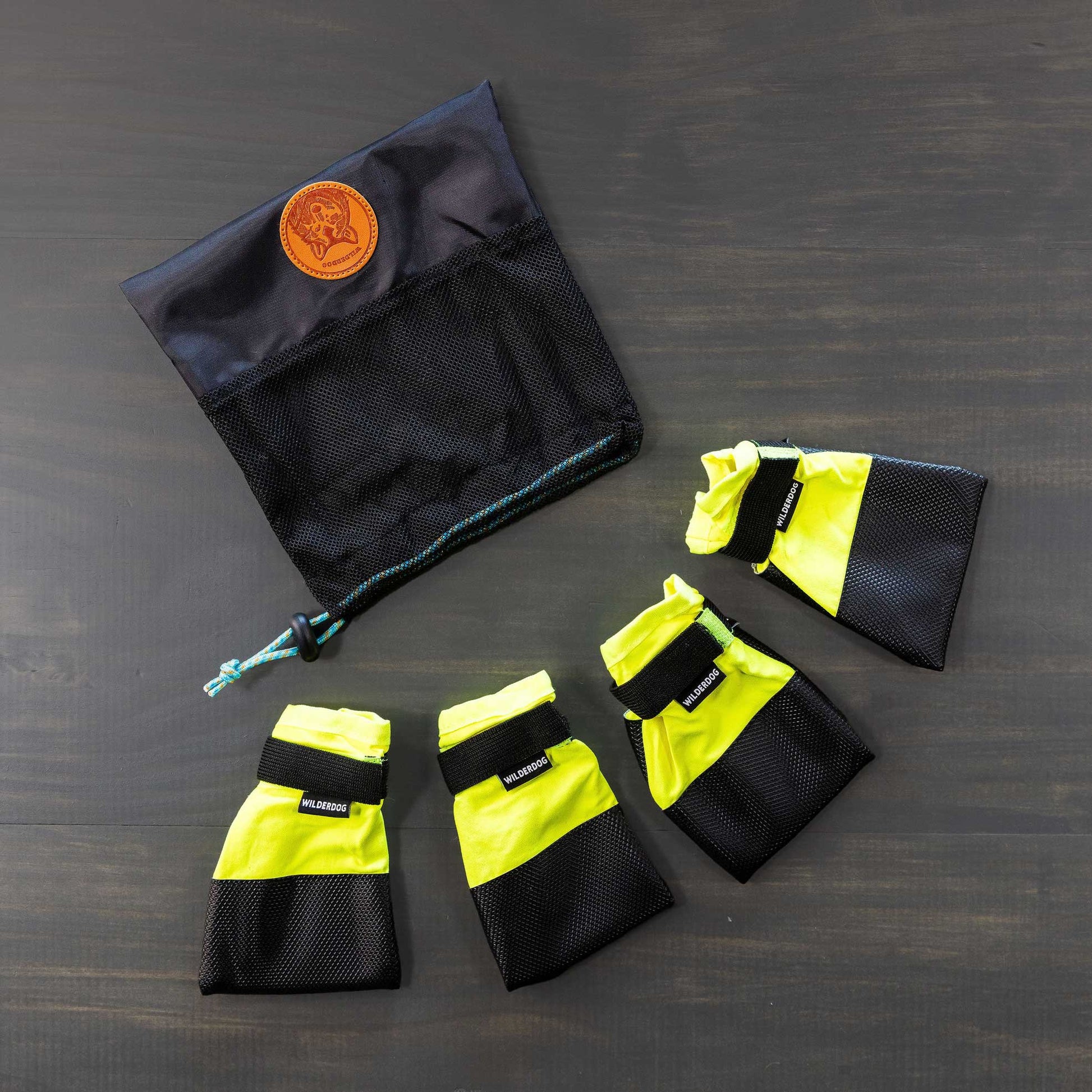 A set of Dog Booties, featuring four yellow and black booties with Velcro straps for excellent paw protection, is arranged in a semi-circle on a gray surface. Positioned above them is a black mesh storage bag with a drawstring and adorned with a brown leather patch.