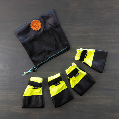 A set of Dog Booties, featuring four yellow and black booties with Velcro straps for excellent paw protection, is arranged in a semi-circle on a gray surface. Positioned above them is a black mesh storage bag with a drawstring and adorned with a brown leather patch.