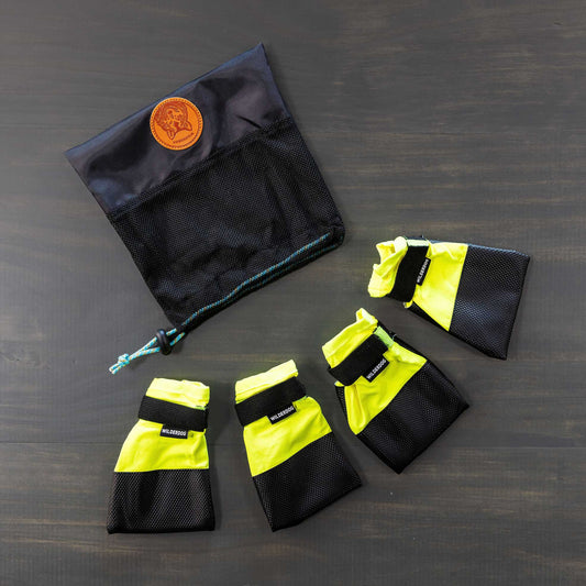 A set of Dog Booties, featuring four yellow and black booties with Velcro straps for excellent paw protection, is arranged in a semi-circle on a gray surface. Positioned above them is a black mesh storage bag with a drawstring and adorned with a brown leather patch.