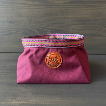A maroon fabric bag with colorful woven trim and a round leather patch embossed with a design makes the perfect carrier for your Backpacking Bowl. Set against a wooden background, it's ideal for any adventure.