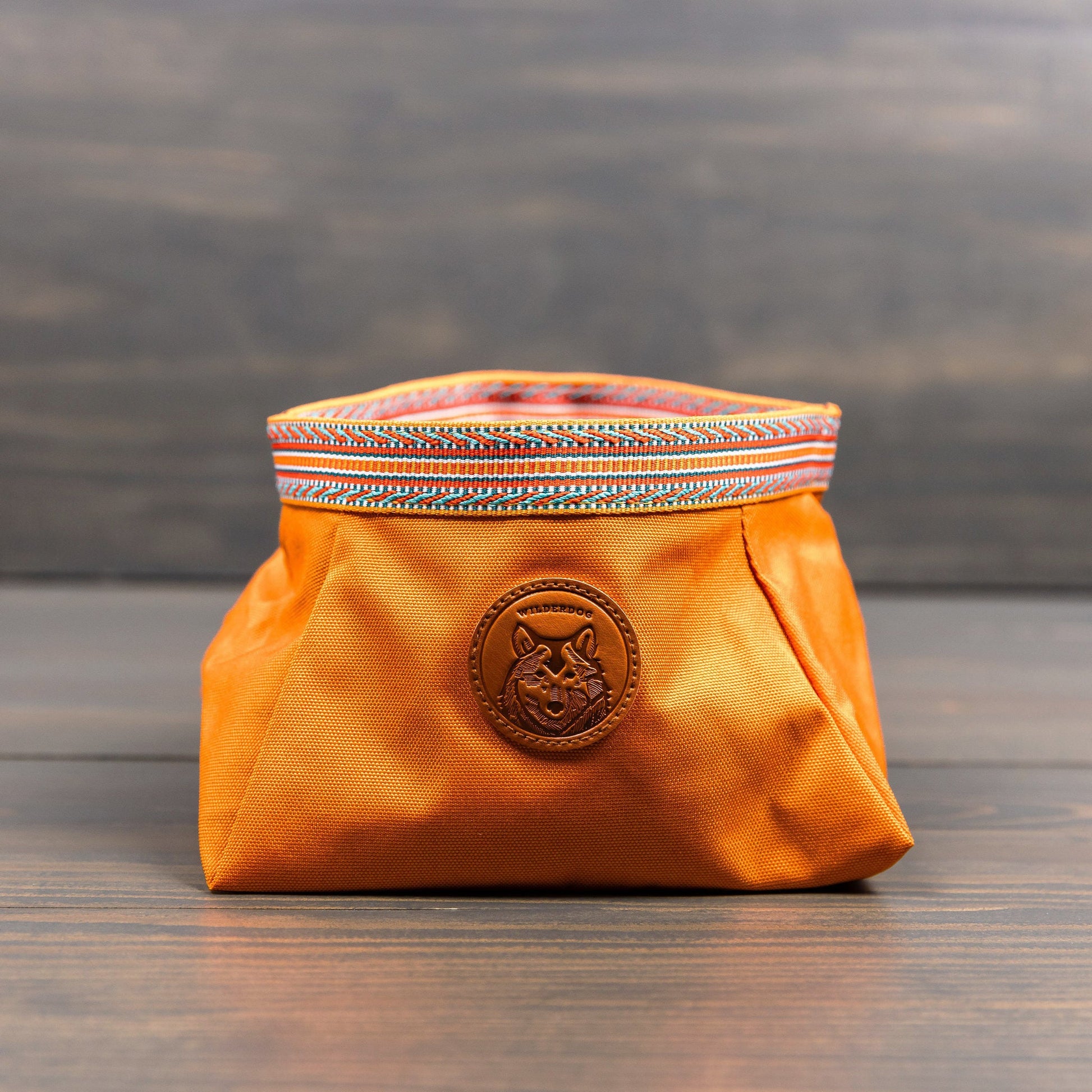 The Backpacking Bowl, a vibrant orange fabric pouch adorned with a colorful woven trim at the top, rests on a wooden surface. It includes a circular patch showcasing a fox design and offers water-resistant qualities, making it ideal for backpacking adventures.