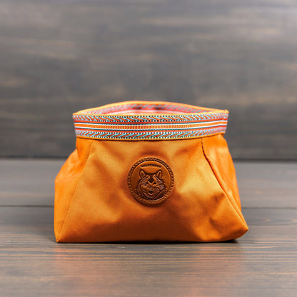The Backpacking Bowl, a vibrant orange fabric pouch adorned with a colorful woven trim at the top, rests on a wooden surface. It includes a circular patch showcasing a fox design and offers water-resistant qualities, making it ideal for backpacking adventures.