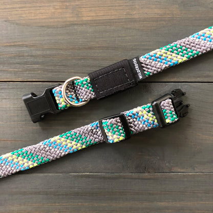 The Alpine Reflective Collar and matching leash set, crafted with the durability of rock climbing rope, displayed on a wooden surface. Featuring a blend of gray, blue, green, and yellow patterns, it includes black plastic clips and a silver D-ring for tags or leash attachment.