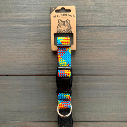The Aprés Collar, a vibrant and durable dog collar adorned with a woven rainbow pattern, is showcased on a wooden surface. It features a black plastic buckle and a metal ring. The packaging displays the brand name "Wilderdog" along with an illustration of a dog on the label.