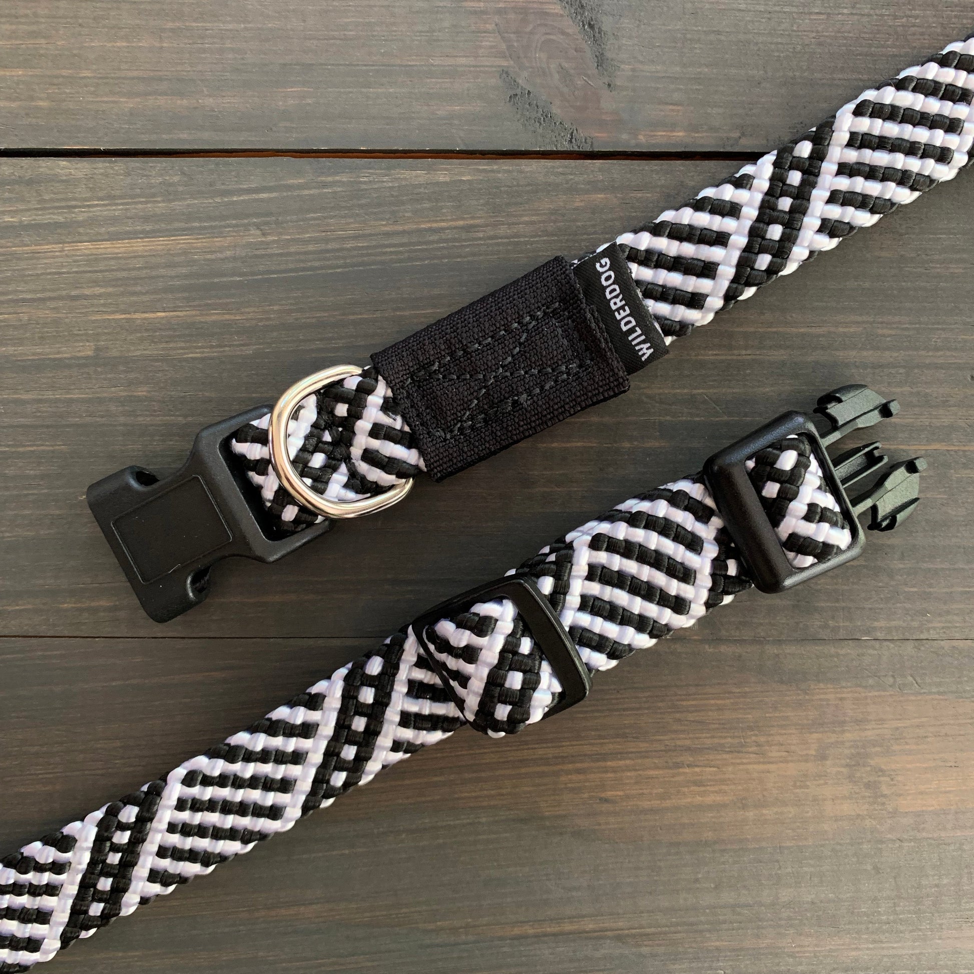 Close-up of two Black and White Collars, resembling black and white woven rock climbing rope, with plastic buckles and metal D-rings, placed on a dark wooden surface. The collars feature interlacing patterns and a small label with text partially visible.