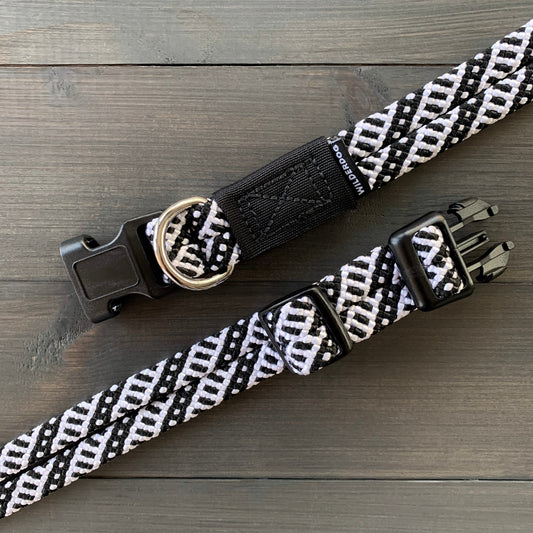 Two Black and White Collars are displayed on a wooden surface. Each heavy-duty dog collar features a sturdy buckle, a metal D-ring for attaching a leash or ID tag, and an adjustable clip. The pattern consists of intertwined black and white threads creating a textured design.